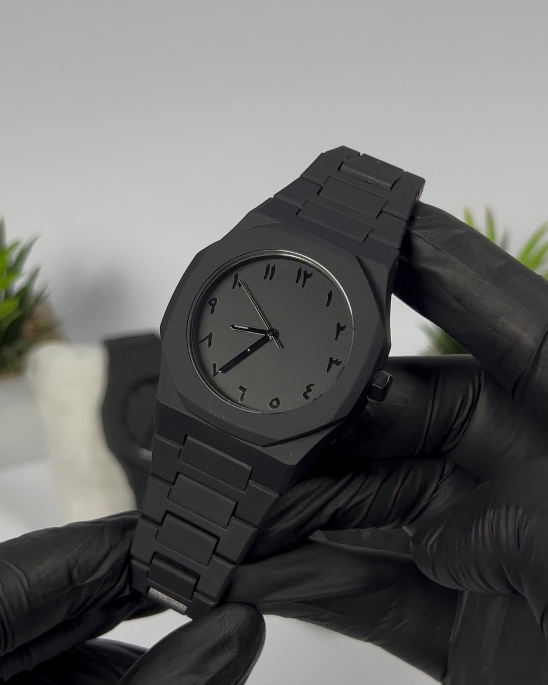 Black wristwatch held by hands wearing black gloves against a neutral background